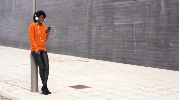 Afro kobieta cieszy się i tańczy podczas słuchania muzyki z telefonu komórkowego i słuchawek na zewnątrz na ulicy. — Wideo stockowe