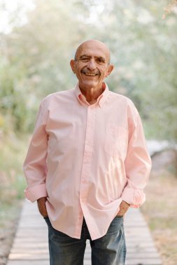 Olgun bir adam kameraya bakıyor ve doğada poz verirken gülümsüyor..