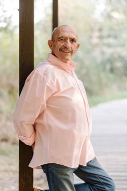 Olgun bir adam kameraya bakıyor ve dışarıda bir sütuna yaslanmış poz verirken gülümsüyor..