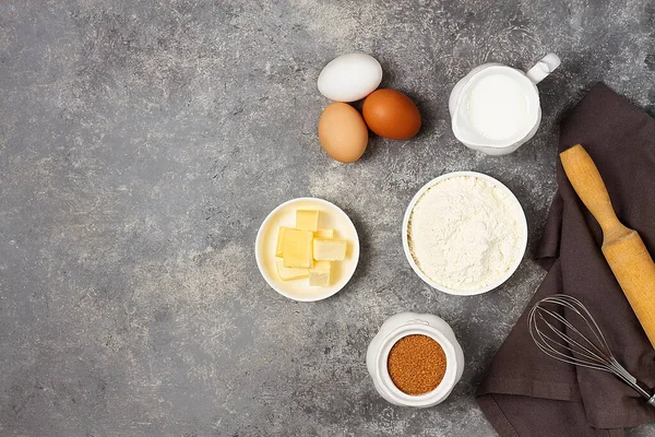 Teigzutaten, Milch, Eier, Mehl und Schneebesen, Butter, brauner Rohrzucker, auf einem grauen Tisch, Draufsicht, Hintergrund, keine Personen, waagerecht — Stockfoto