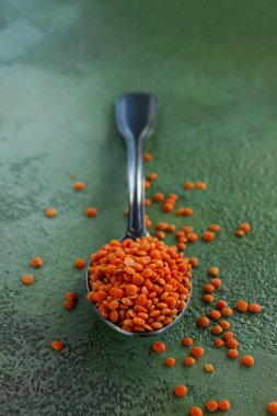 Siyah kaşık parlak turuncu mercimeklerden oluşan bir tepecikte, desenli yeşil bir yüzeye dökülen mercimekler, canlı renk kontrastını ve dokusunu vurgular..