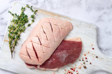 Taze çiğ ördek göğsü, ahşap bir kesim tahtasına yayılmış, beyaz mermer bir yüzey üzerinde otlar ve baharatlarla,