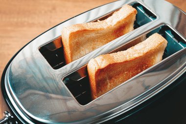 İki dilim ekmek, sıcak bir mutfaktaki tezgahın üzerinde duran parlak metal bir tost makinesinde kızartılıyor. Yumuşak ışık, sıcak bir kahvaltı atmosferini akla getiriyor..