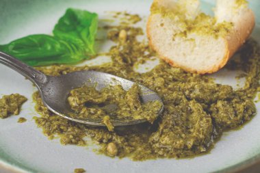 Tabakta taze fesleğenden yapılmış bir pesto sosu, bir dilim ekmek ve tabakta bir kaşıkla birlikte. Pesto sosu kremalı ve yeşil görünüyor. Ekmek de kısmen yenmiş..