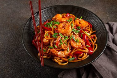 Kırmızı dolmalık biber ve yeşil soğanlı udon kızartmalı karides eriştesi, siyah bir kasede servis ediliyor, çubuklarla servis ediliyor, koyu arka plan...,