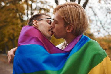 Romantik eşcinsel çift dışarıda sarılıyor, öpüşüyor ve el ele tutuşuyor. Parkta LGBT gurur bayrağı taşıyan iki yakışıklı adam..