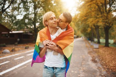 Romantik eşcinsel çift dışarıda sarılıyor, öpüşüyor ve el ele tutuşuyor. Parkta LGBT gurur bayrağı taşıyan iki yakışıklı adam..