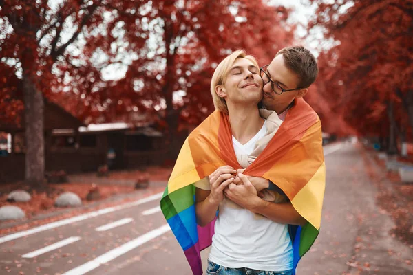 Romantik eşcinsel çift dışarıda sarılıyor, öpüşüyor ve el ele tutuşuyor. Parkta LGBT gurur bayrağı taşıyan iki yakışıklı adam..