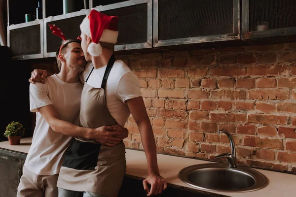 Evdeki sevecen eşcinsel çift. Noel Baba şapkasıyla mutfakta sarılıp öpüşen iki yakışıklı adam. Noel havası. LGBT kavramı.