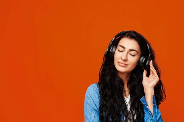 Cheerful girl listening to music. Stylish teenager enjoying of a new self playlist music over orange studio background. Free space