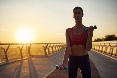 Dambıllarla sabah egzersizleri, karantina koşullarında. Sağlıklı yaşam tarzı kavramı. Gün doğumunda dambıllarla egzersiz. Güneş kameraya doğru parlıyor.