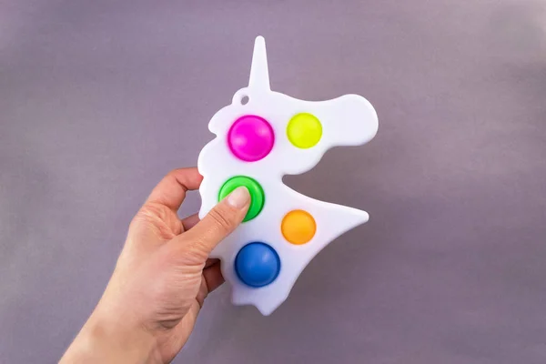 Ljusa färgglada leksak Simple Dimple enhörning. Populär anti-stress leksak på en grå bakgrund i händerna på en kvinna Stockbild