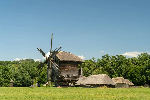 Antiguo molino de viento y casas rurales tradicionales ucranianas medievales en un prado verde. Cielo azul con nubes blancas. — Foto de Stock