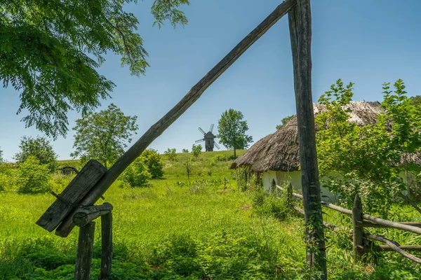 Antiguo molino de viento y casas rurales tradicionales ucranianas medievales en un prado verde. Cielo azul con nubes blancas. — Foto de Stock