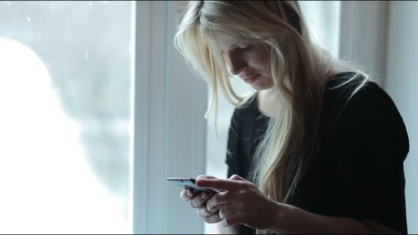 Menina bonita perto da janela com um smartphone, conversando — Vídeo de Stock