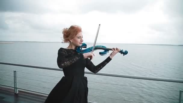 Giovane donna in abito nero che suona il violino sul molo sullo sfondo di una vista incredibilmente bella del fiume. Steadicam shot — Video Stock