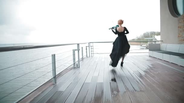 Молодая женщина в черном платье играет на скрипке на причале на фоне удивительно красивого вида на реку. Стедикам-шот — стоковое видео