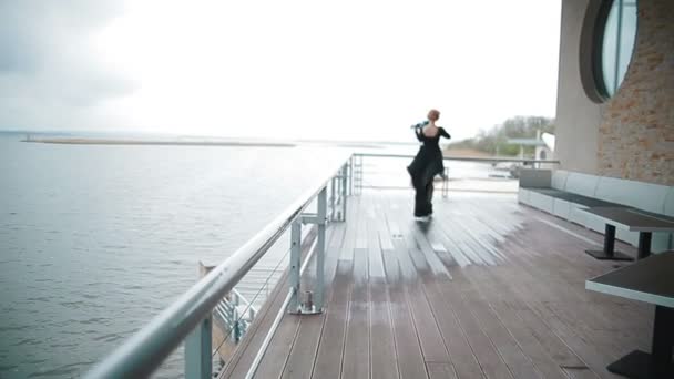 Giovane donna in abito nero che suona il violino sul molo sullo sfondo di una vista incredibilmente bella del fiume. Steadicam shot — Video Stock