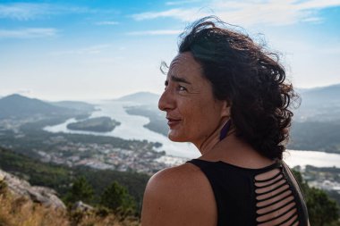 Genç, gülümseyen, kıvırcık saçlı, arka planda bir nehrin ağzında kanyonu olan beyaz bir kadının portresi..