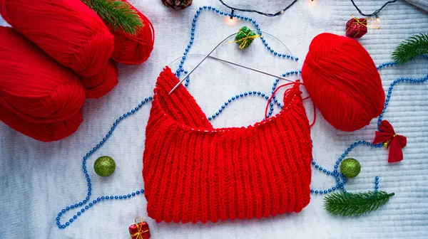 Вязание красных рождественских свитеров на фоне новогоднего декора. Готовимся к празднованию Рождества и Нового года. Вязание вручную. Шерстяные шарики из красной нитки. Рукоделие. — стоковое фото
