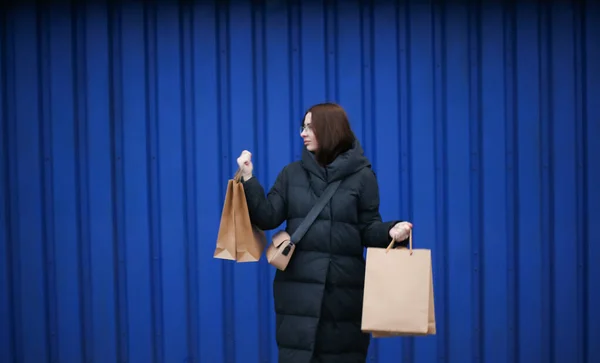 Jeune femme en vêtements d'hiver avec des sacs en papier avec des achats sur fond bleu — Photo