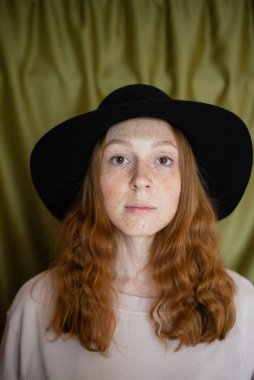 Siyah şapkalı kızıl saçlı kız kameraya bakıyor.
