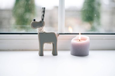 Kış manzaralı bir pencerenin arka planında mum ve oyuncak tahta geyik.