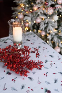 Mum, cam bir şamdanda. Arka planda üzerinde ışıklar olan bir Noel ağacı var. Masadaki şenlikli masa örtüsü.