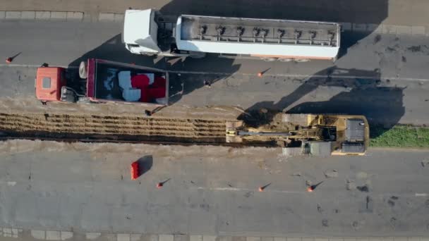 Gran excavadora de orugas cava terreno entre dos carriles de autopista y grandes parachoques de carretera de descarga de camiones — Vídeo de stock