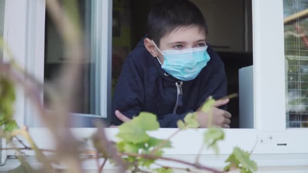 Βαρετό παιδί σε καραντίνα στο σπίτι. Αγόρι με προστατευτική ιατρική μάσκα για την πρόληψη του ιού της στέψης κοιτάζει έξω από το παράθυρο — Αρχείο Βίντεο
