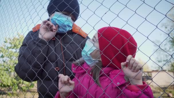 Menino e menina em máscara médica protetora para prevenção de coronavírus atrás de rede no parque infantil — Vídeo de Stock