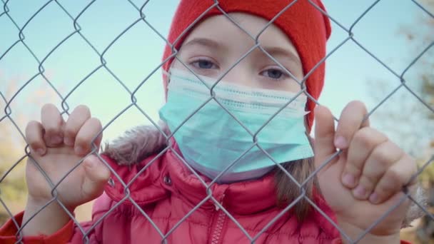 Sad girl in protective medical mask for coronavirus prevention standing behind netting during quarantine of epidemic — Stock Video