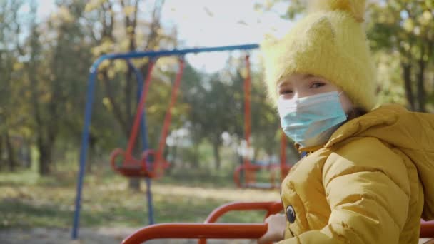 Retrato de niña en máscara médica se ve tristemente en el parque infantil durante la epidemia de virus en todo el mundo — Vídeos de Stock