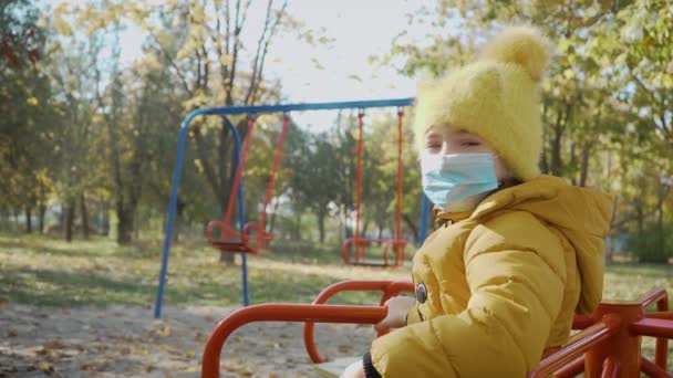 Маленька дівчинка в медичній захисній масці сидить сама без своїх друзів під час карантину коронавірусу — стокове відео