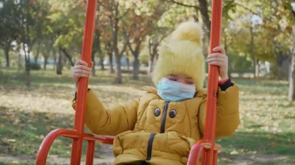 Niña con máscara protectora médica montada en columpio sola sin sus amigos durante la cuarentena del coronavirus — Vídeos de Stock