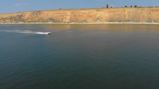 Vista lateral de grande lancha branca com pessoas em coletes salva-vidas se movendo rápido ao longo da praia do mar com tendas camping — Vídeo de Stock