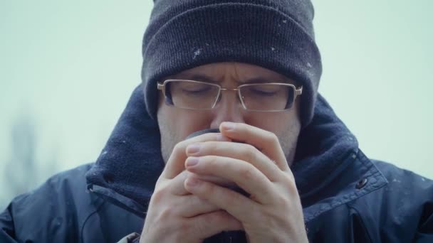 El hombre sostiene la taza de vapor caliente y la bebe en la tormenta de nieve. Taza de camping de metal de hojalata con café o té fresco especial de la mañana — Vídeos de Stock