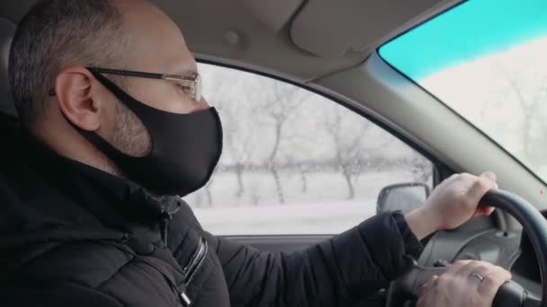 Вид сбоку на белого человека в противовирусной маске, который ездит в машине, используя и держа руль зимой — стоковое видео