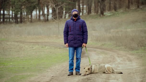 Человек в черной защитной маске для профилактики коронавируса и очках стоит на улице с собакой на поводке в лесу зимой во время пандемии — стоковое видео