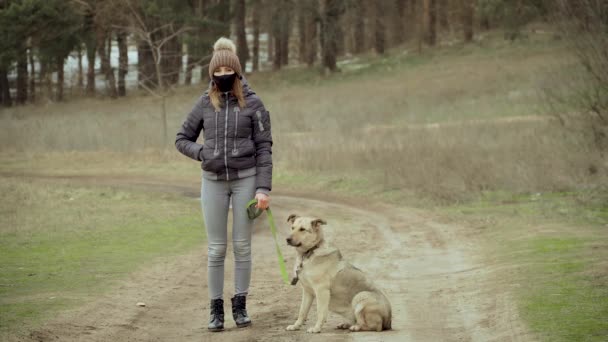 Женщина в черной защитной маске для предотвращения коронавируса стоит на улице с собакой на поводке в лесу зимой — стоковое видео
