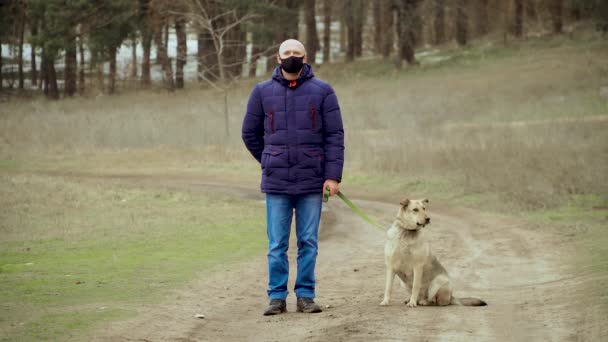 Muž v černé ochranné anti korona virus tvář maska nehybný stát venku se psem na vodítku v blízkém lese v zimě během vlastní izolace na karanténě — Stock video