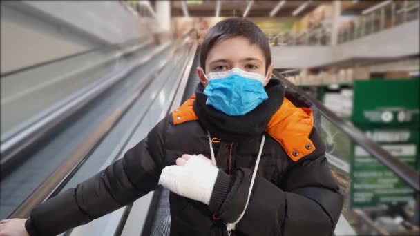 Мальчик в защитной медицинской маске для предотвращения коронавируса со сломанной рукой в гипсе едет на эскалаторе в торговом центре или торговом центре — стоковое видео