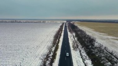 Drone, kırsal asfalt yolda araba ve kamyon sürüşü videosu çekiyor. Kışın tarlalar arasında her iki tarafında da kar var.
