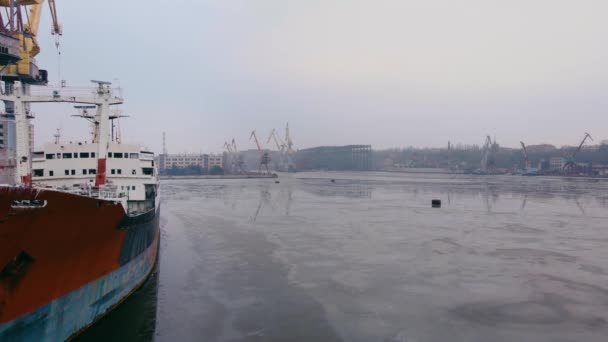 Lucht panning in de buurt van grote roestige schip afgemeerd in vrachthaven in de buurt van grote multi tonnage kranen in smeltend ijs op bevroren water — Stockvideo