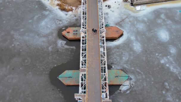 Pandangan atas ke bawah pada orang-orang yang tidak dikenal berjalan di atas jembatan pejalan kaki baja melalui sungai dengan es yang mencair di musim dingin — Stok Video