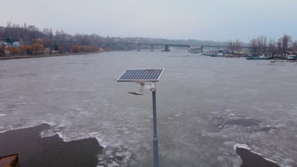 Маленькая солнечная панель на стальном пешеходном мосту через реку с тающим льдом в холодное время — стоковое видео