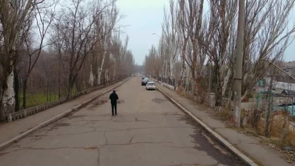 Grim view άνθρωπος περπατά κατά μήκος του παλιού δρόμου με ραγισμένη άσφαλτο και λίγα σταθμευμένα αυτοκίνητα στο πεζοδρόμιο και φυλλώδη δέντρα στο πάρκο — Αρχείο Βίντεο