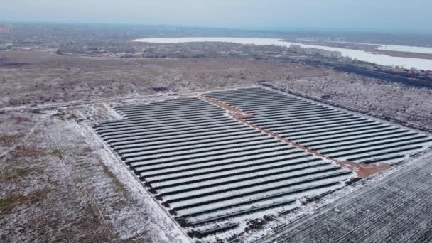 Panneaux solaires photovoltaïques debout dans un champ neigeux sous un ciel hivernal lourd près d'une petite ville — Video