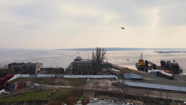 从空中俯瞰直升机在造船厂上空飞驰的景象，它配备着巨大的运货起重机，很少有船只站在水面上，浮冰融化 — 图库视频影像