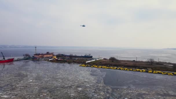 Large transport helicopter flies over transport harbor with artificial bay and moored cargo ships in melting ice — ストック動画